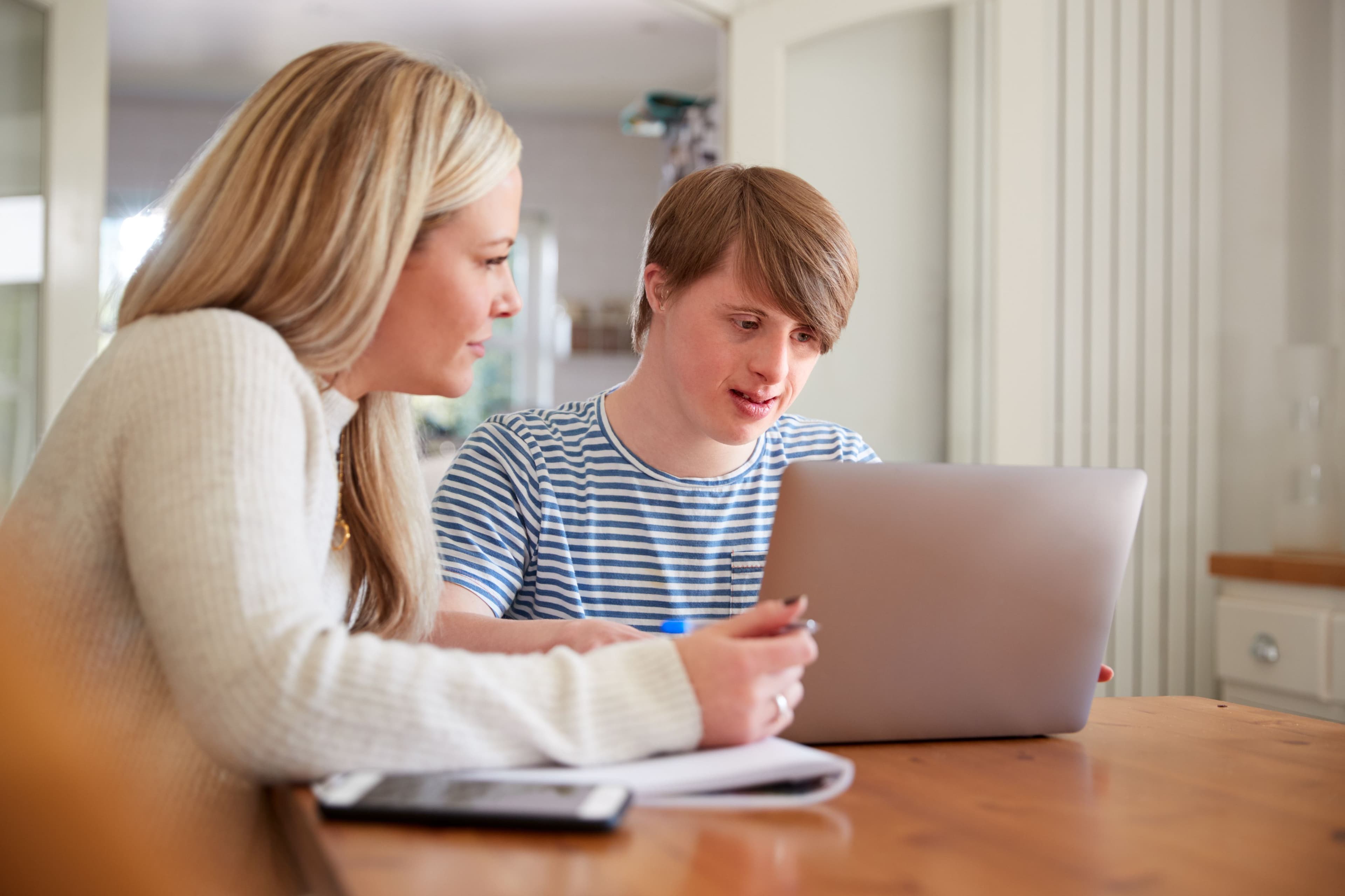 A young man with Down syndrome and a woman sit at a table, looking at a laptop.